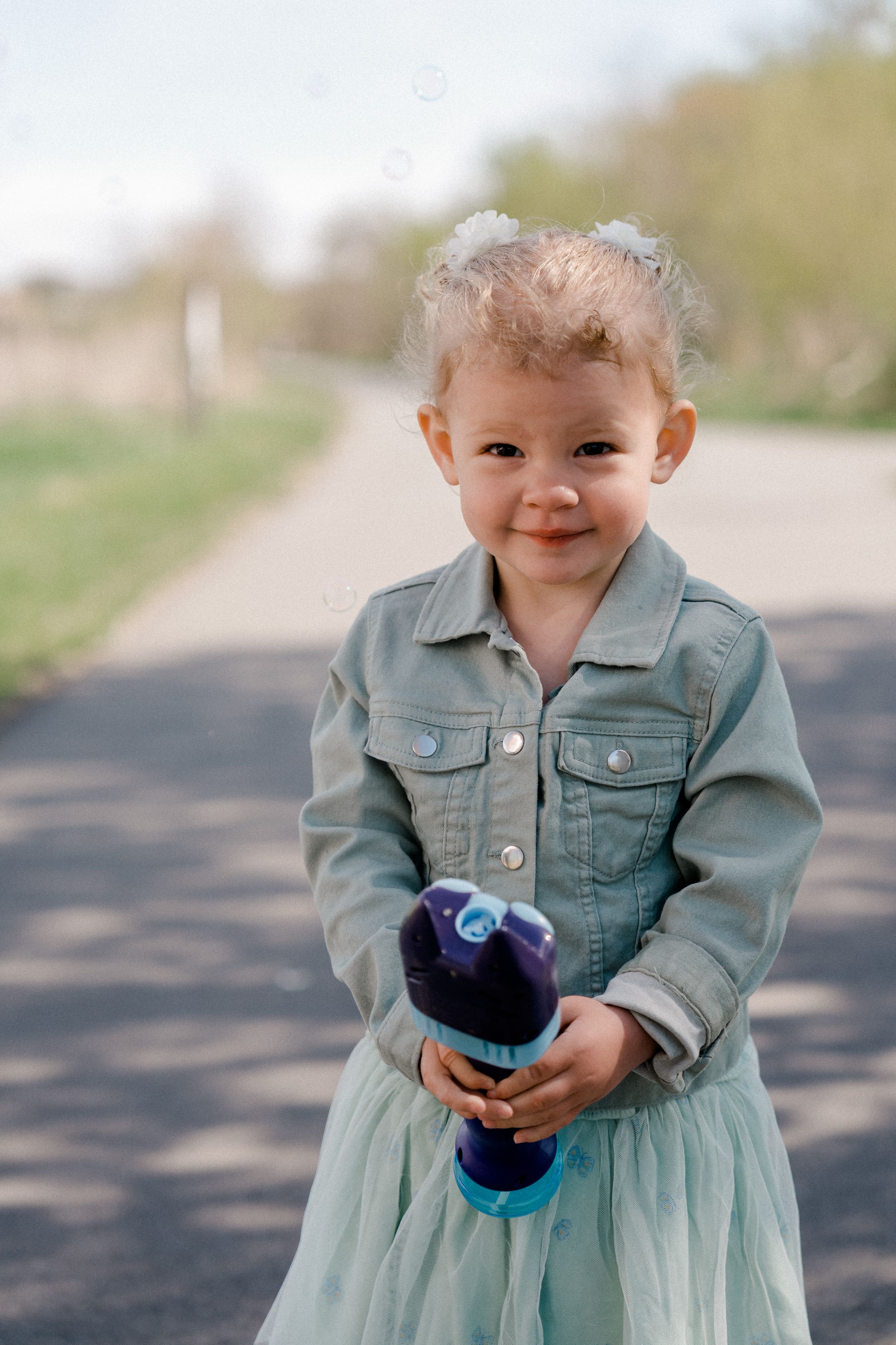 Small child wearing light denim jacket and tulle skirt holds purple water bottle while standing on path outdoors.
