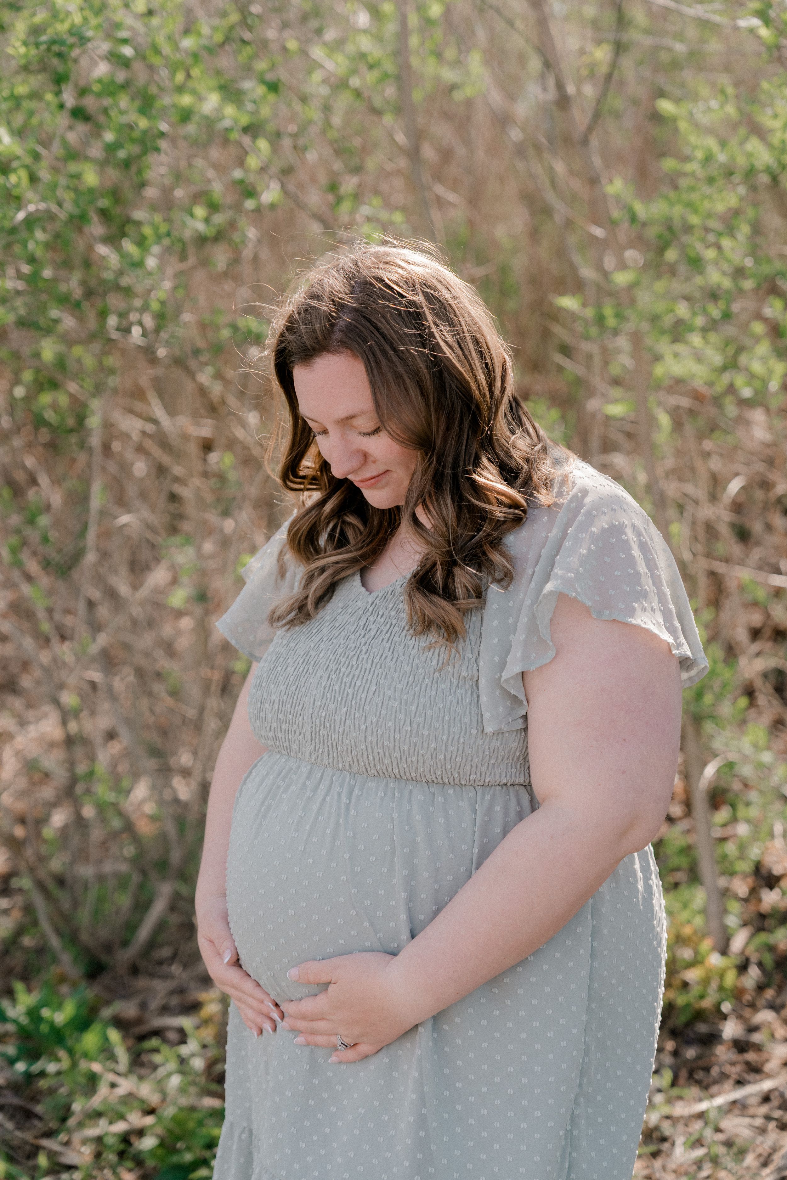 A pregnant woman in a light gray dress stands outdoors with her hand gently resting on her baby bump.