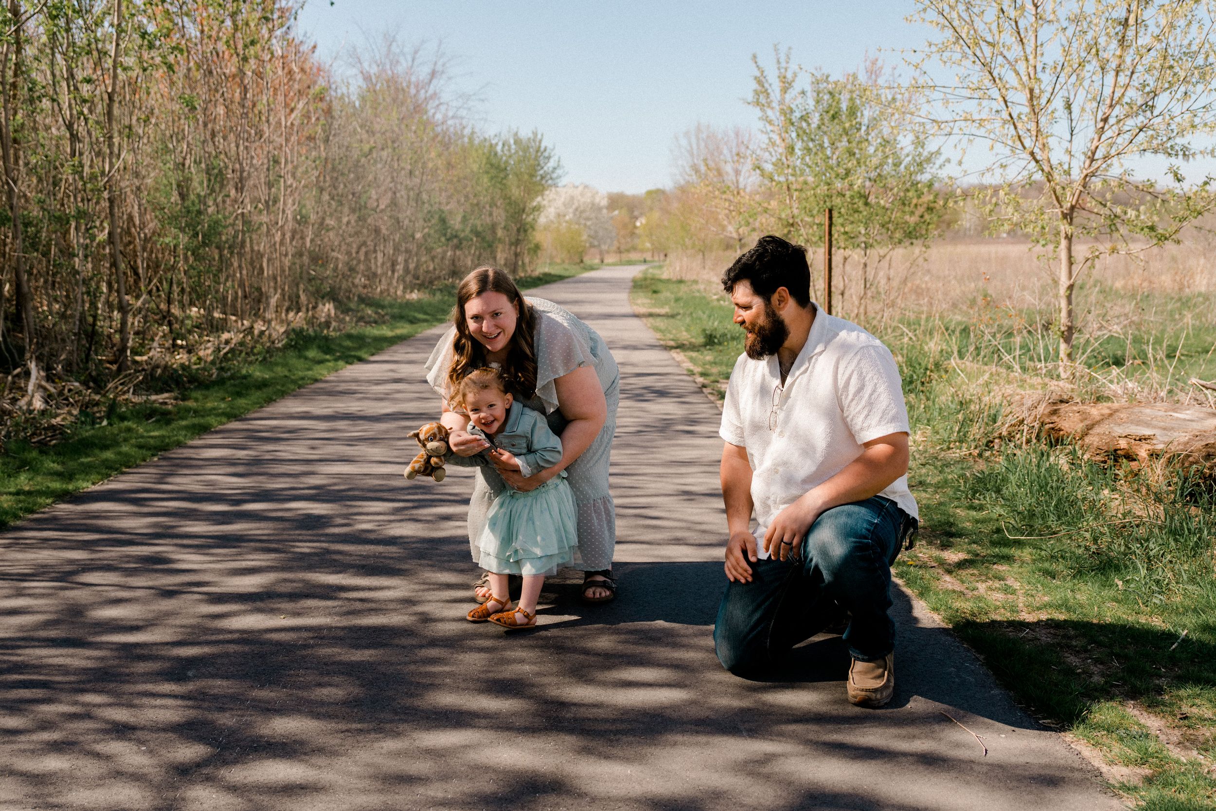 Two people kneeling on a paved nature trail with their small dog on a sunny spring day.
