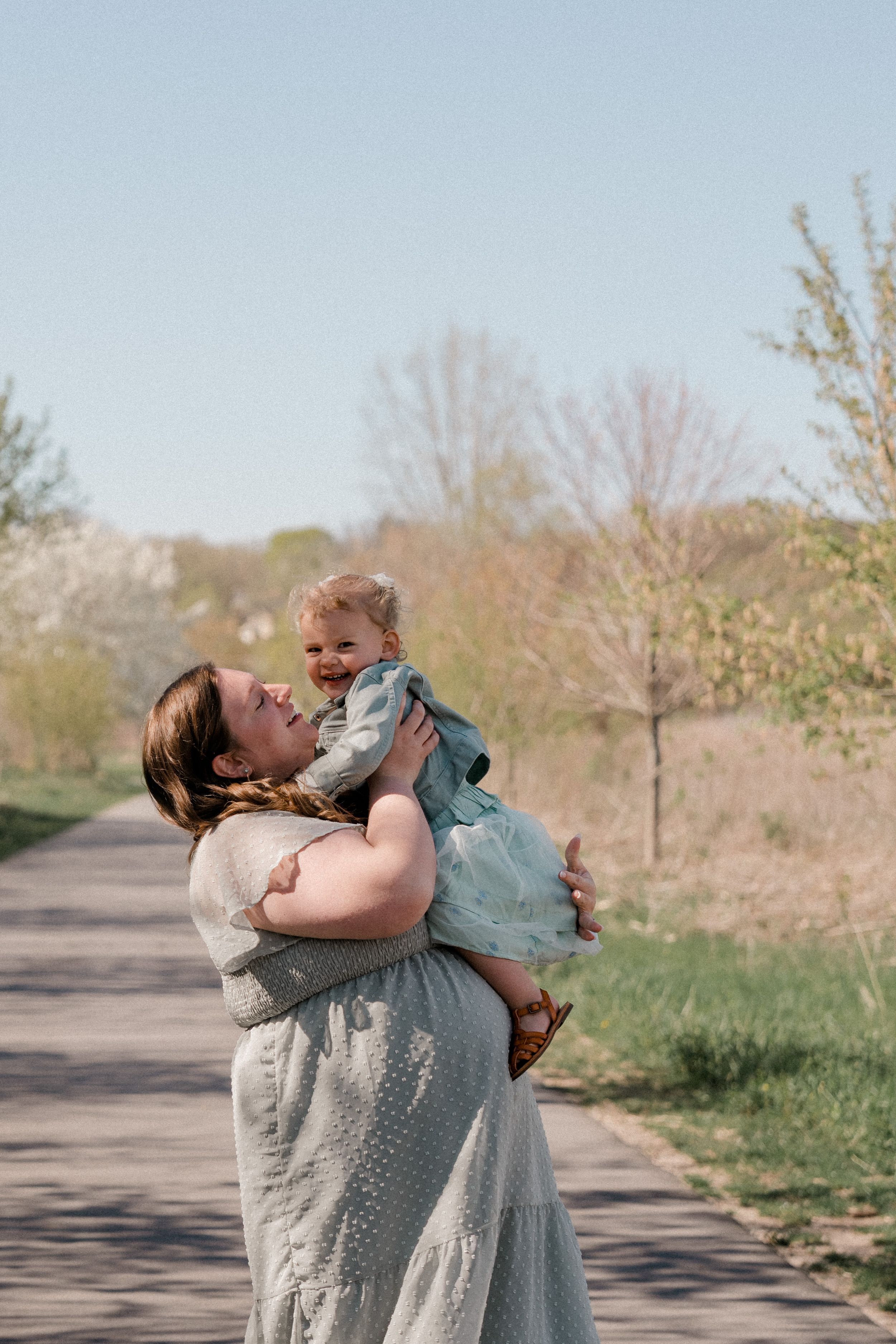 A joyful moment shared between a woman in a grey dress and a small child on a tree-lined pathway in spring.