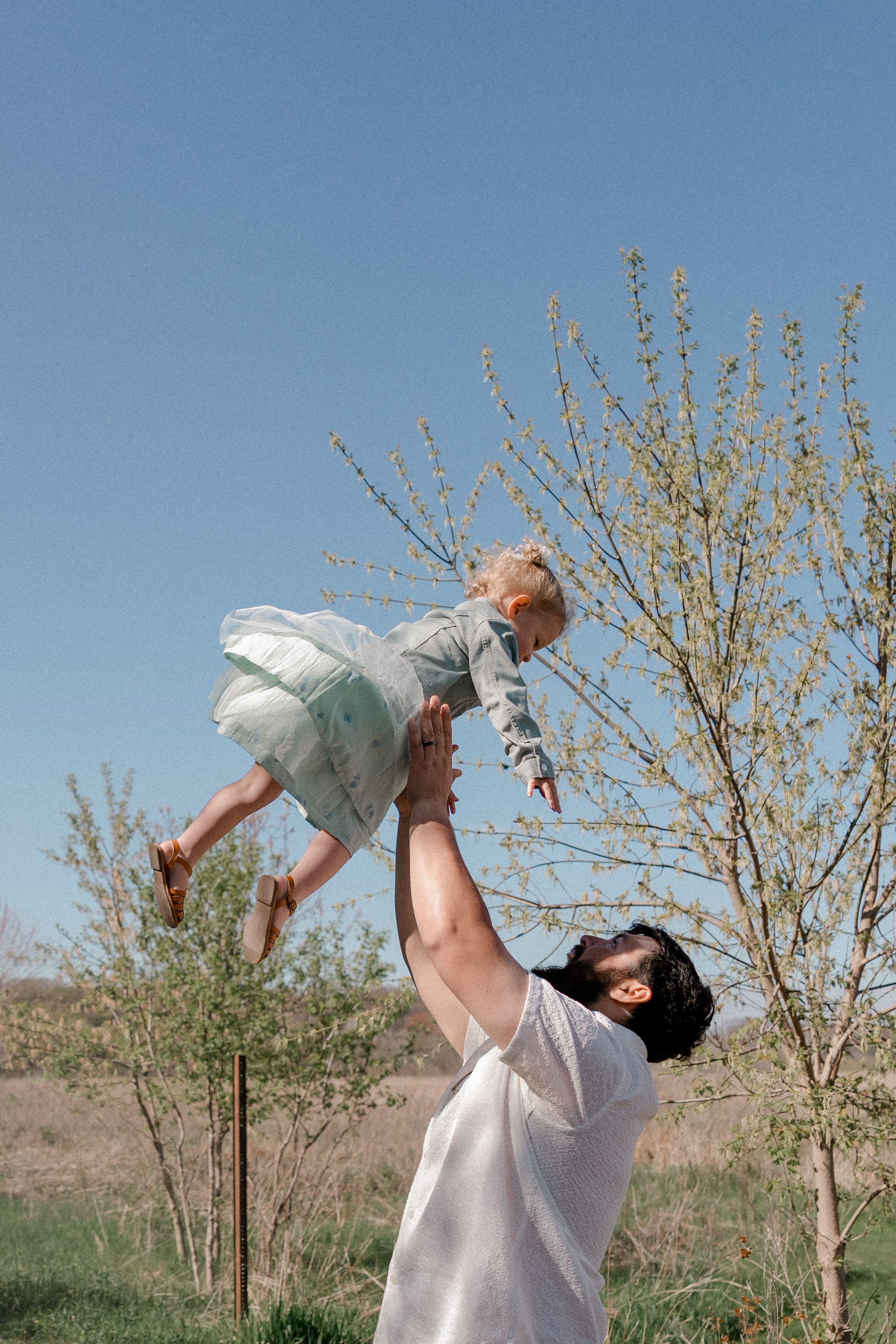 A person in white lifts and balances a child wearing light clothing against a blue sky with budding trees in background.