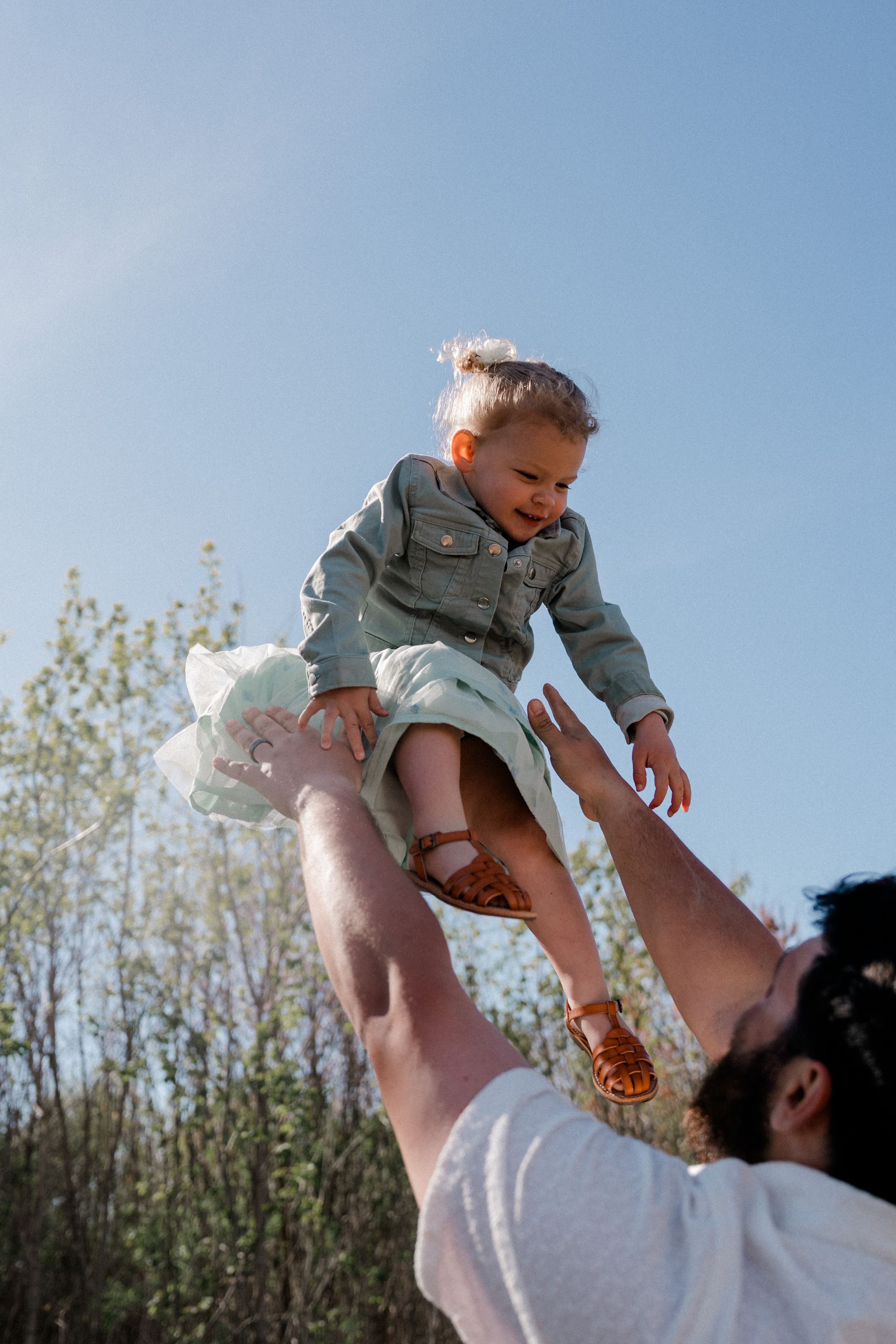 Toddler in denim jacket being lifted up high against a blue sky and spring foliage on a sunny day.