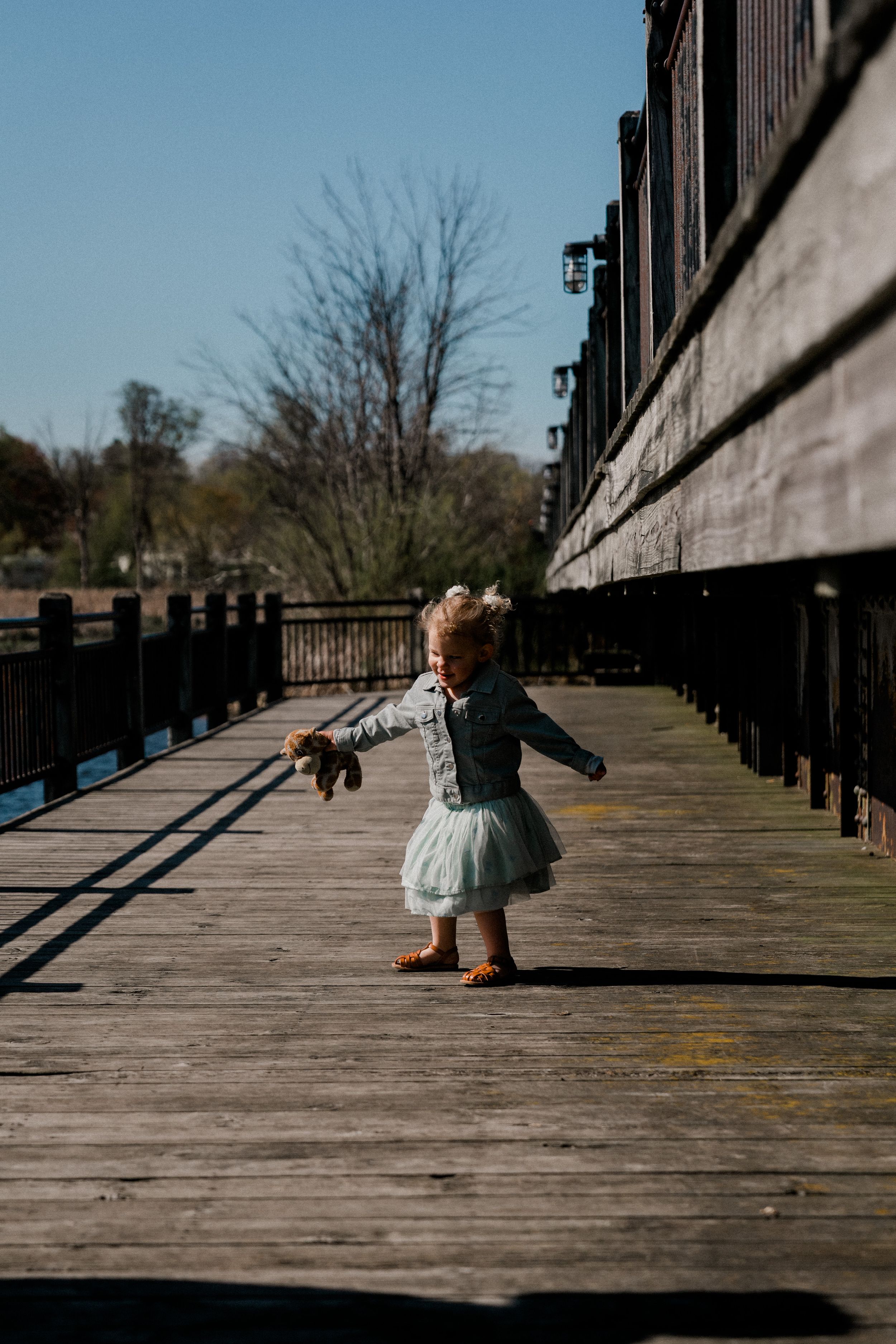 Small child in a tulle dress twirls on a wooden boardwalk on a sunny day with long shadows.