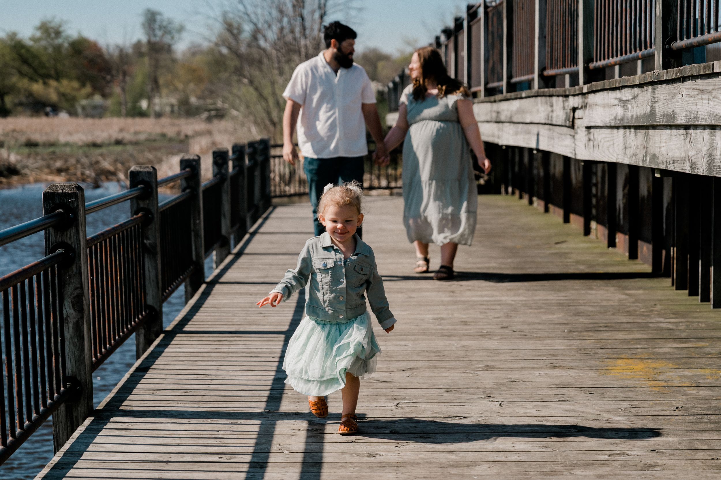 Young child in teal dress runs joyfully ahead on wooden boardwalk while two people follow behind on sunny day.