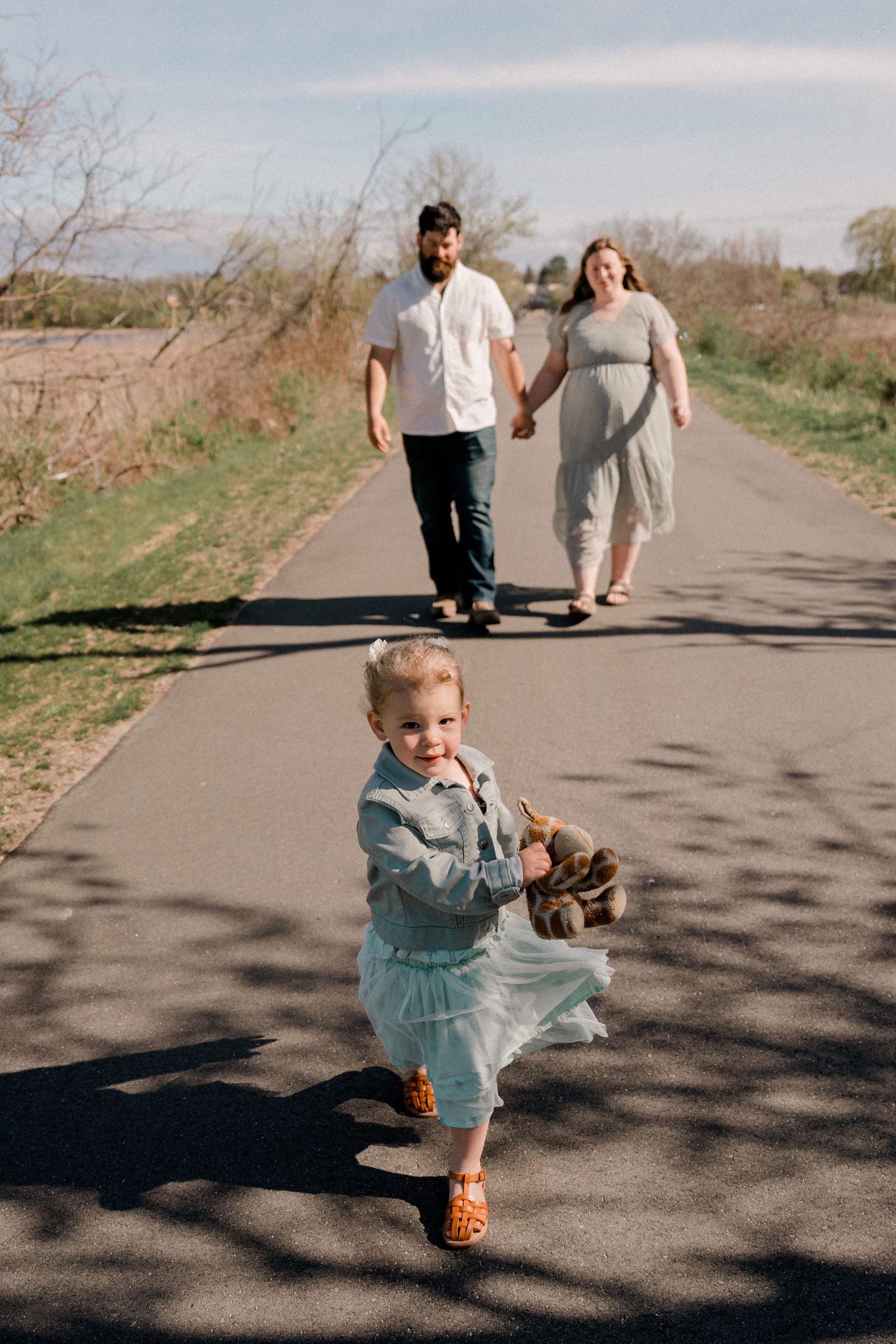 Two people walk hand in hand on a paved path while a child in a tulle dress runs ahead on a sunny day.