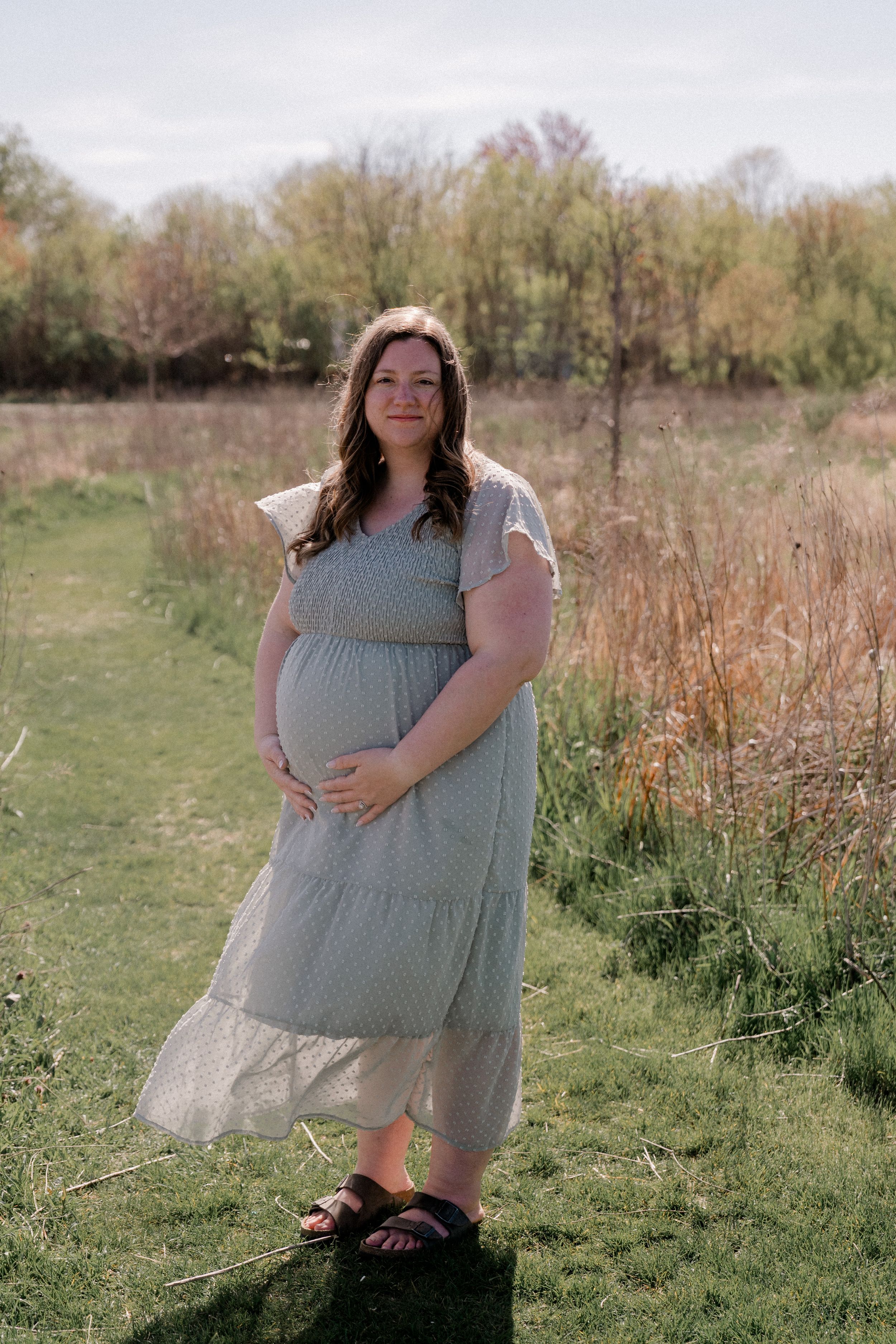 A pregnant person in a tiered grey dress stands in a sunlit meadow surrounded by tall grass and trees.