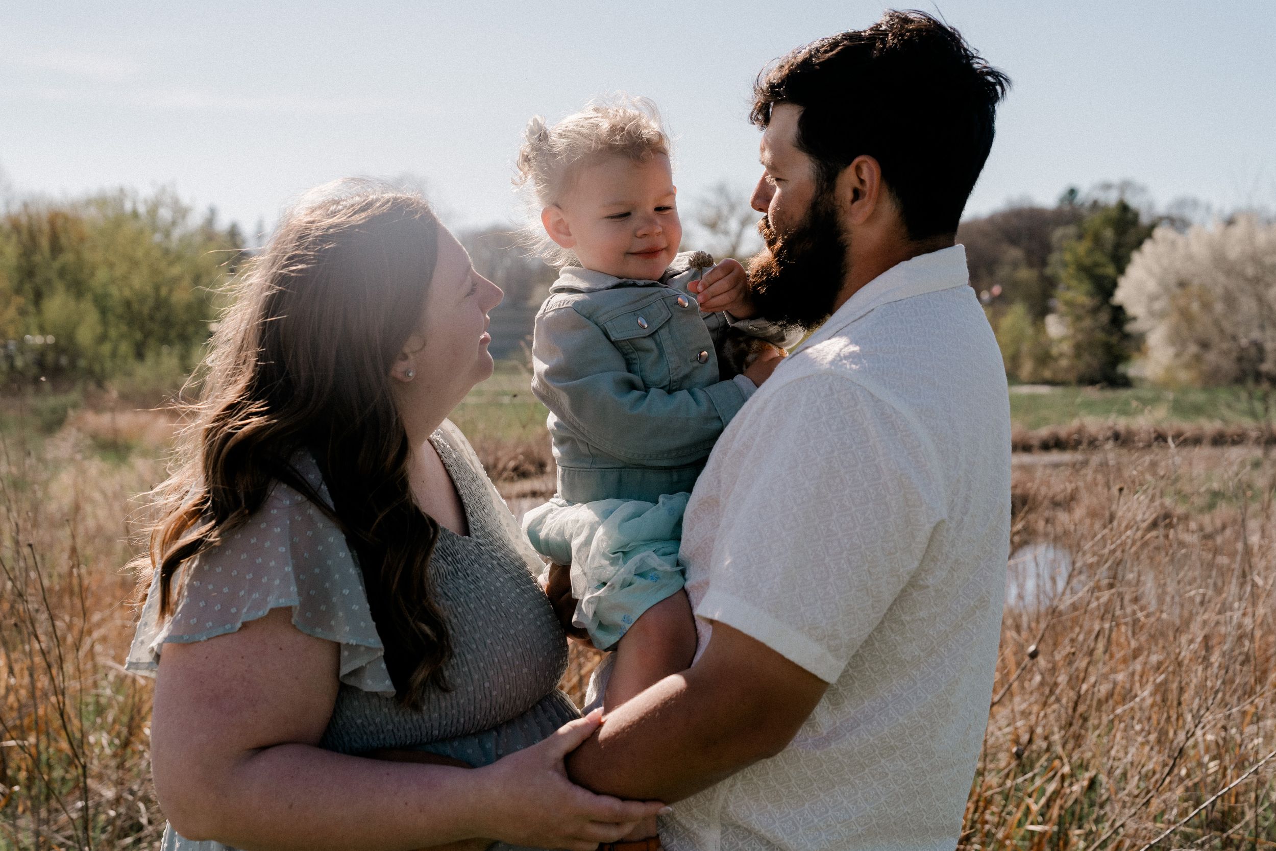 Three people stand together in a golden field during sunset as they share a tender family moment.