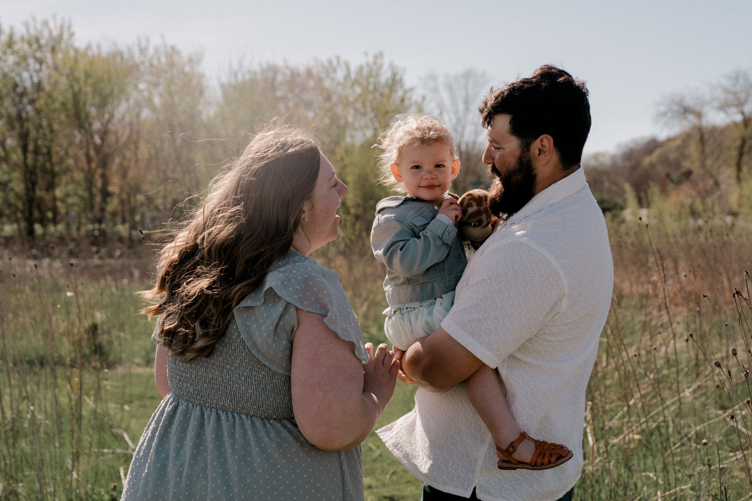 A couple stands together in a grassy field during golden hour holding their young child.