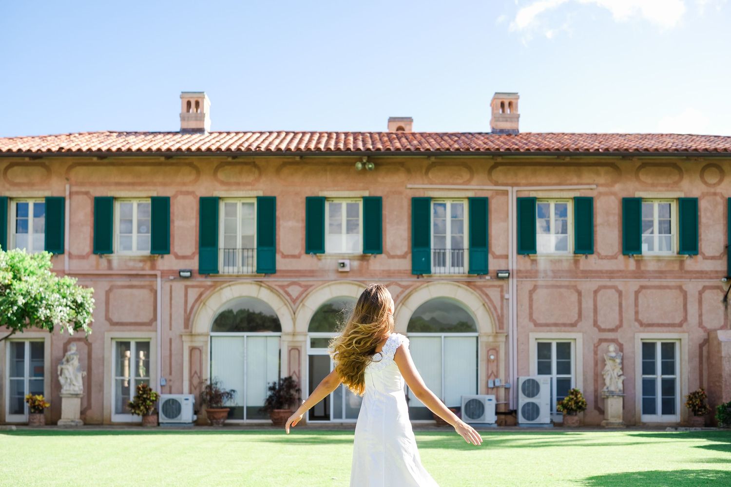 A scenic villa in Mediterranean style with arched doorways, green shutters, and pink facade stands behind a manicured lawn.Oahu Family Photo