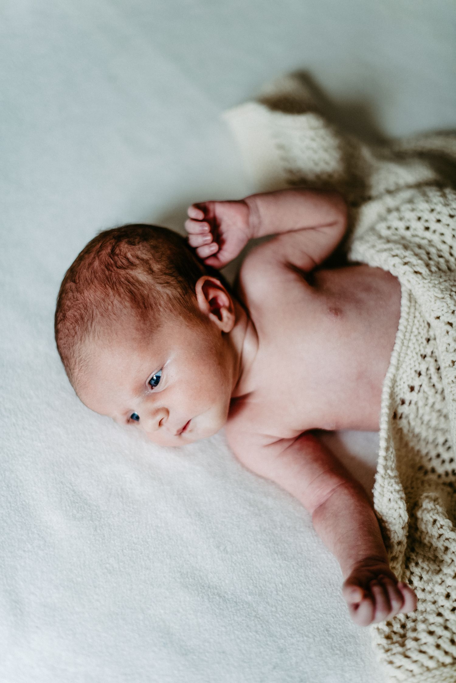Neugeborenes Baby liegt auf einer weißen Decke, teilweise mit einer gestrickten Decke bedeckt, und schaut zur Seite.