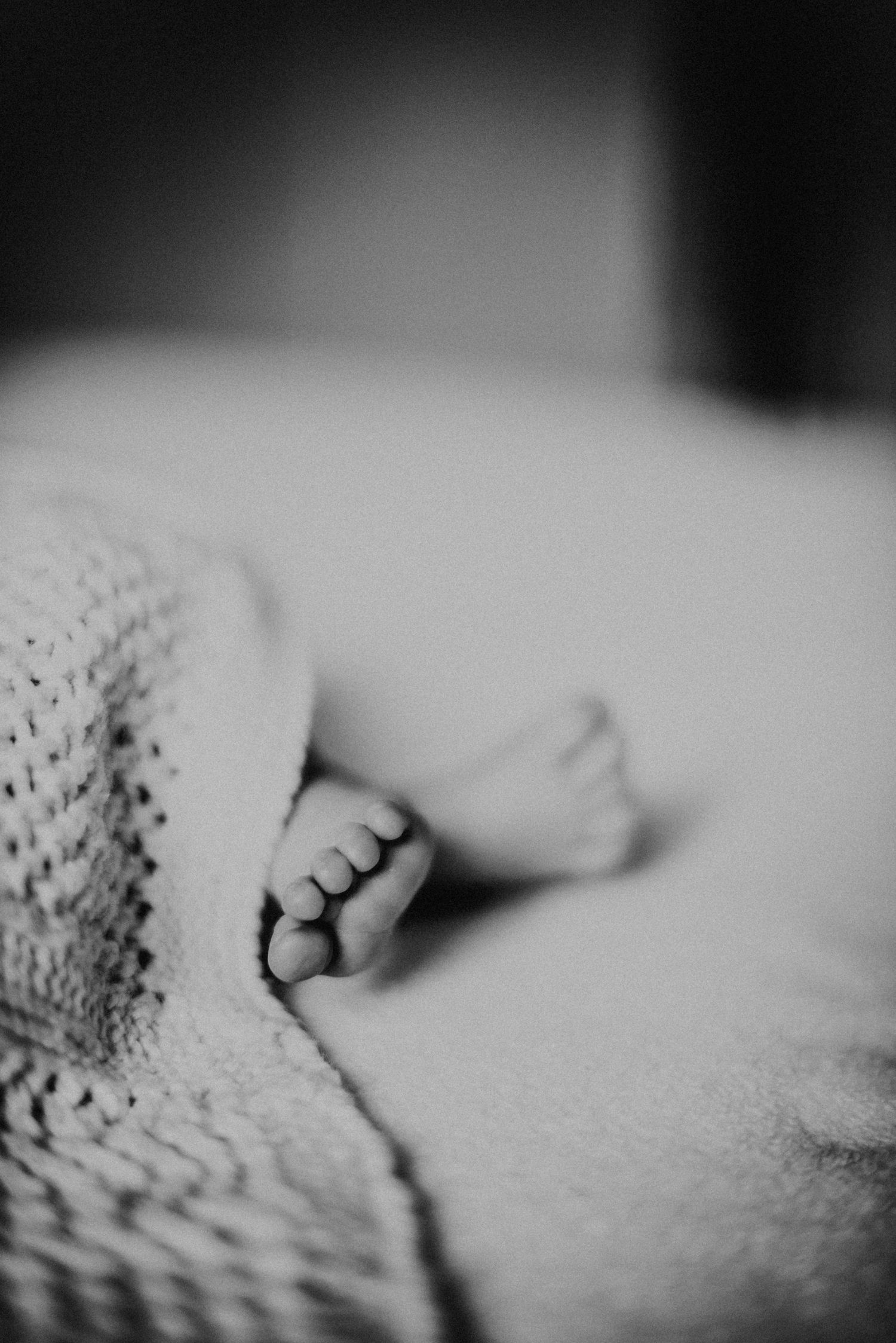 Schwarz-weiß-Foto von Babyfüßen, die unter einer gestrickten Decke hervorschauen. Die Decke hat ein Lochmuster.
