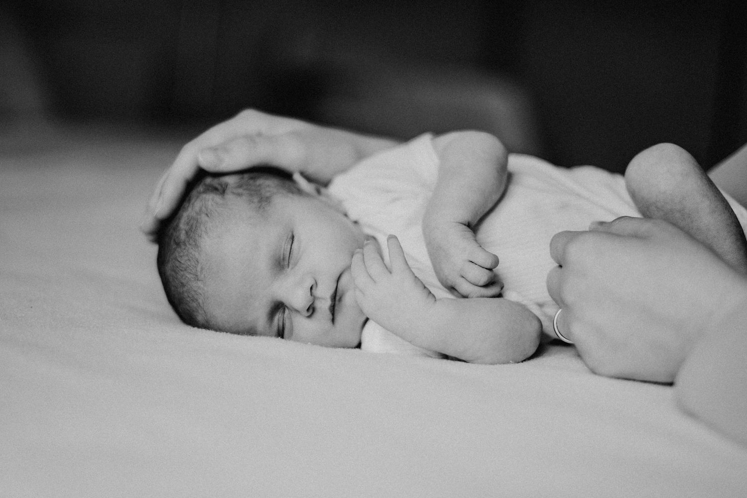 Schwarz-weiß-Foto eines schlafenden Babys in einem weißen Strampler, das sanft von einer Hand auf einem Bett gehalten wird.