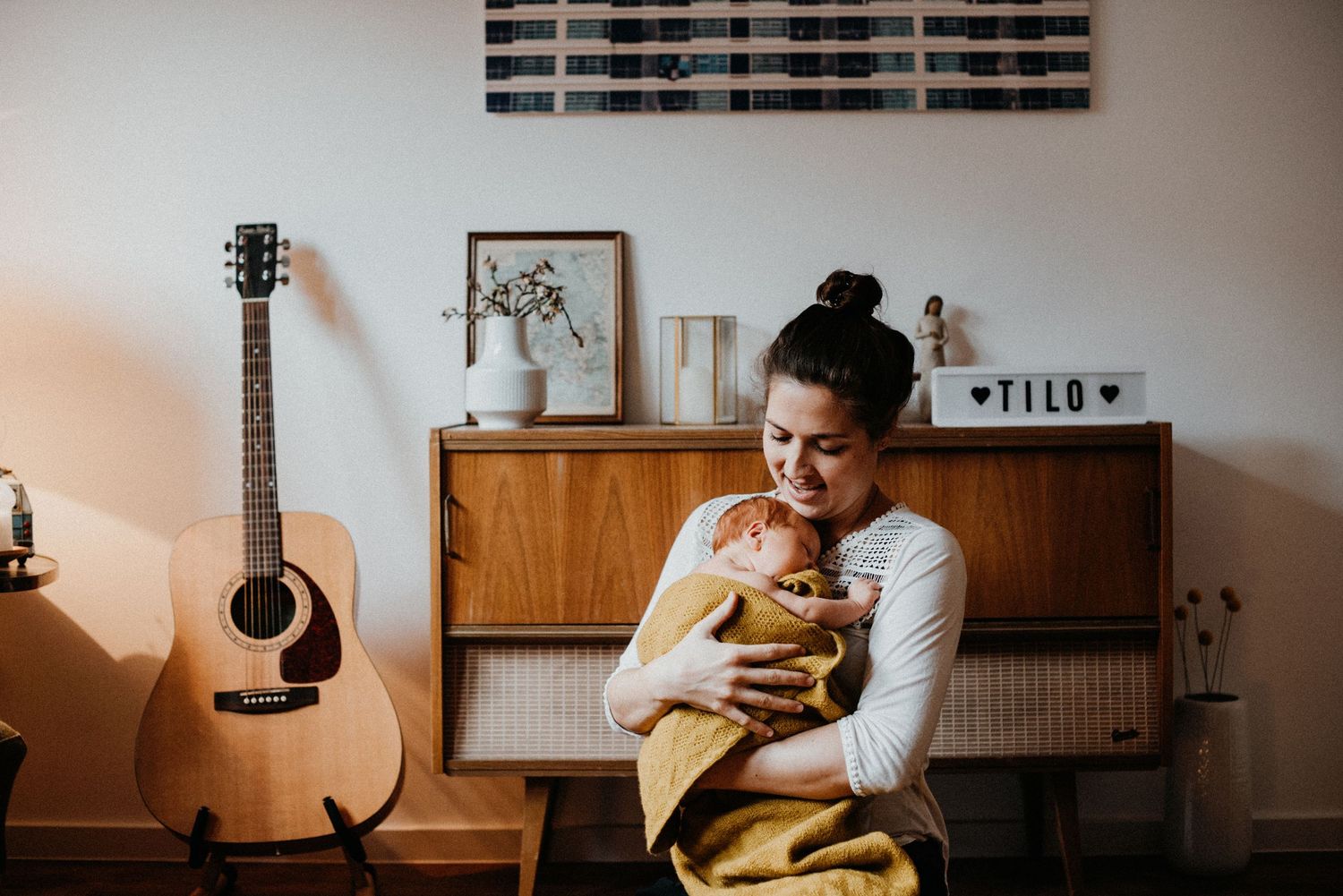 Frau hält ein Baby in einem Wohnzimmer mit Gitarre und Dekorationen. Ein Schild mit dem Namen "Tilo" steht auf dem Regal.