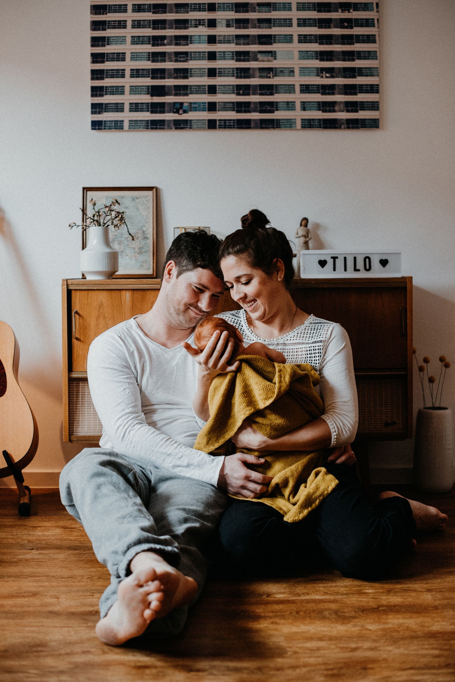 Mama und Papa halten ihr Neugeborenes in den Armen. Sie sitzen auf dem Boden des Wohnzimmers vor einem kleinen Schrank.