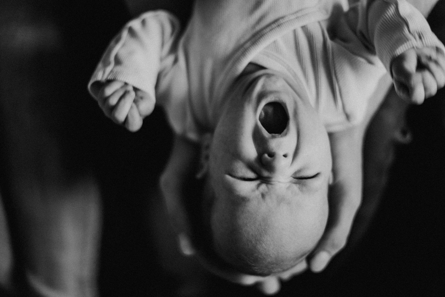 Ein Baby wird kopfüber gehalten und gähnt mit weit geöffnetem Mund. Die Szene ist in Schwarz-Weiß aufgenommen.