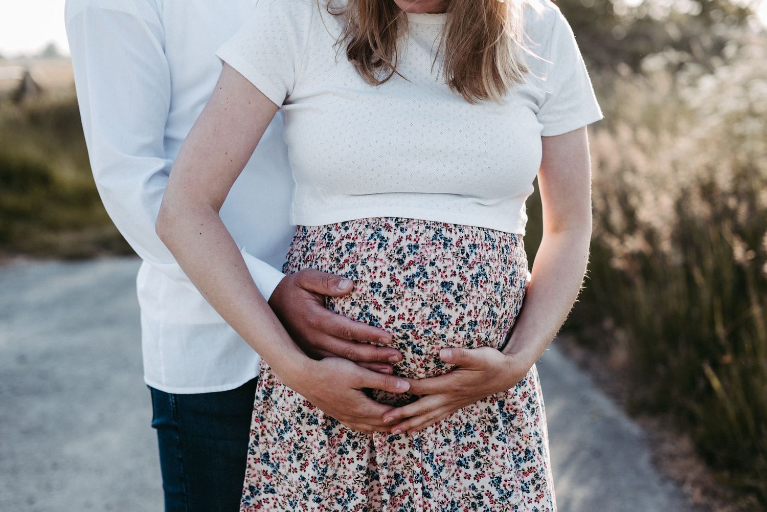 Ein Paar steht auf einem Weg. Die Frau trägt ein geblümtes Kleid und der Mann hält sanft ihren Babybauch.