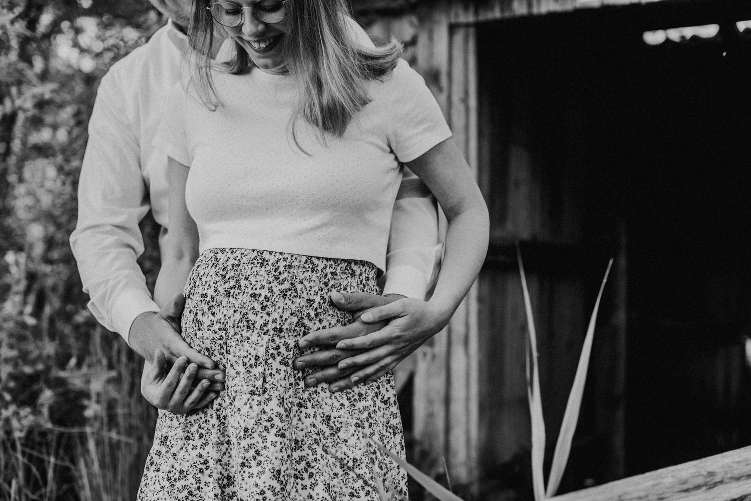 Ein Paar steht auf einem Weg. Die Frau trägt ein geblümtes Kleid und der Mann hält sanft ihren Babybauch.