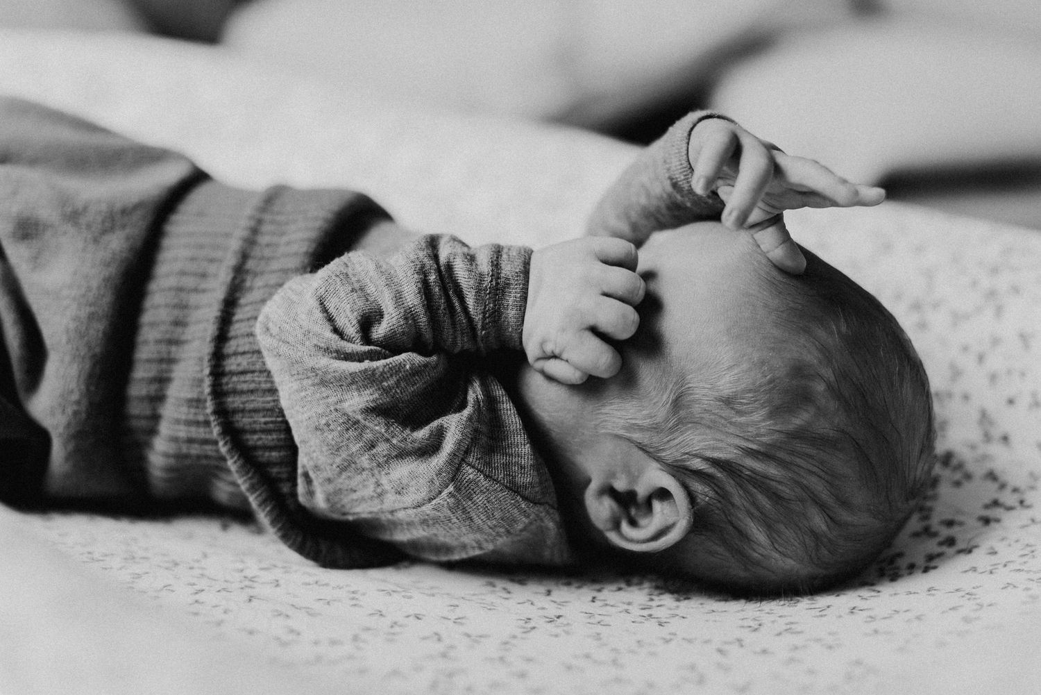 Ein Baby liegt auf dem Rücken und hält seine Hände vor das Gesicht. Es trägt einen Strampler und liegt auf einer gemusterten Decke.