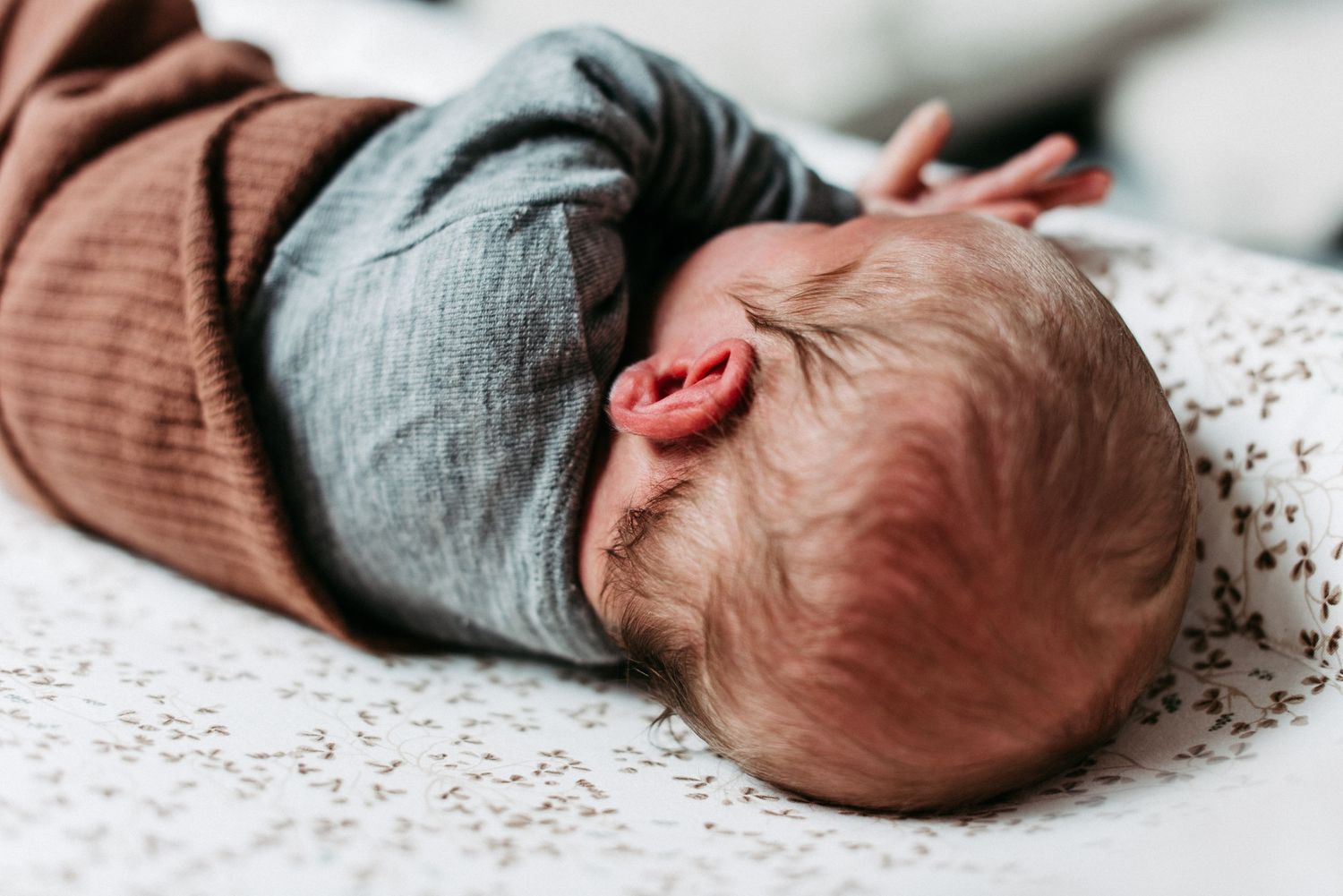 Ein schlafendes Baby liegt auf einer gemusterten Decke, in einen braunen Strampler gekleidet, mit dem Kopf zur Seite gedreht.