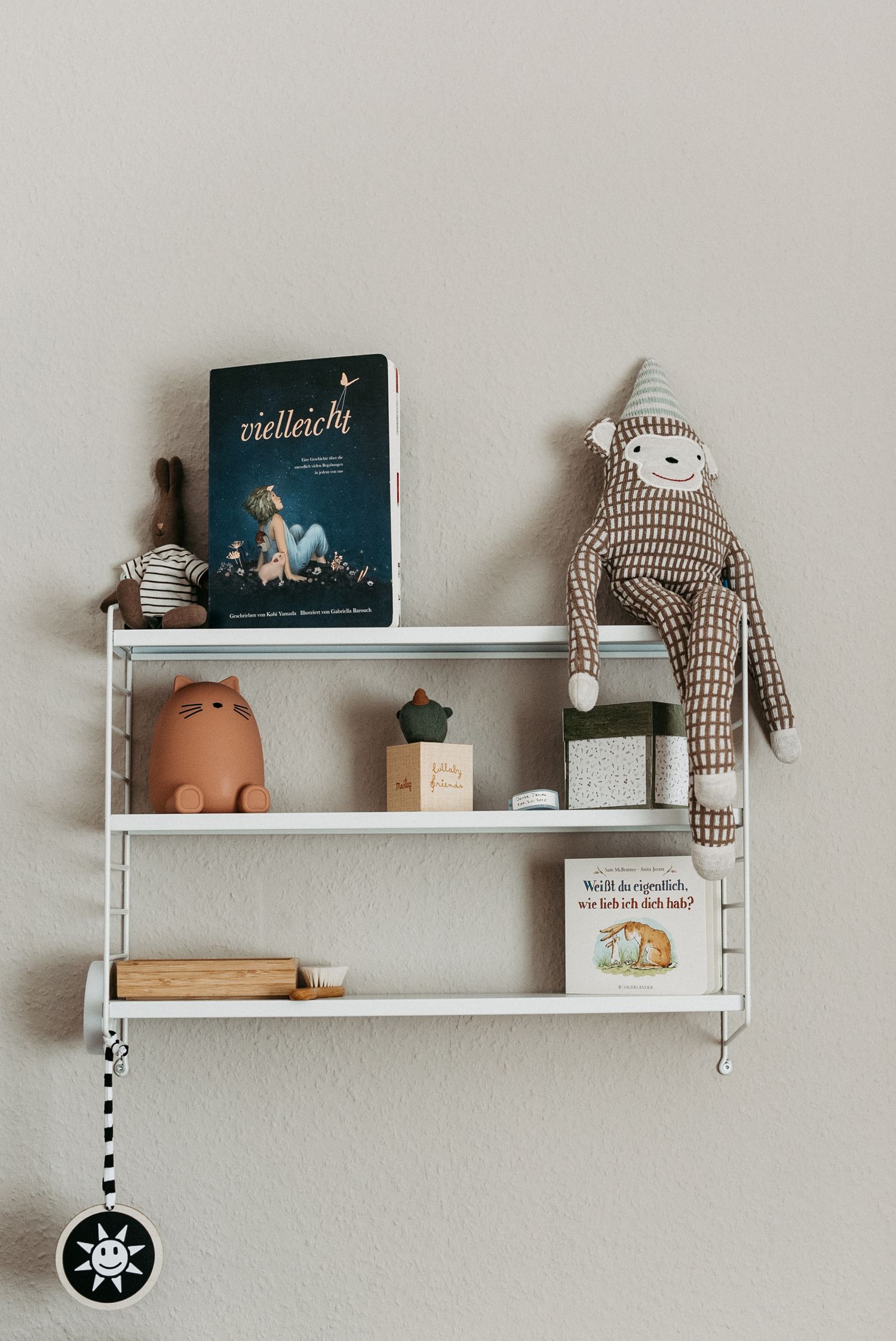 Ein weißes Wandregal mit einem Buch, Stofftier, Kerzen und Dekorationen. Das Regal hängt an einer beigen Wand.