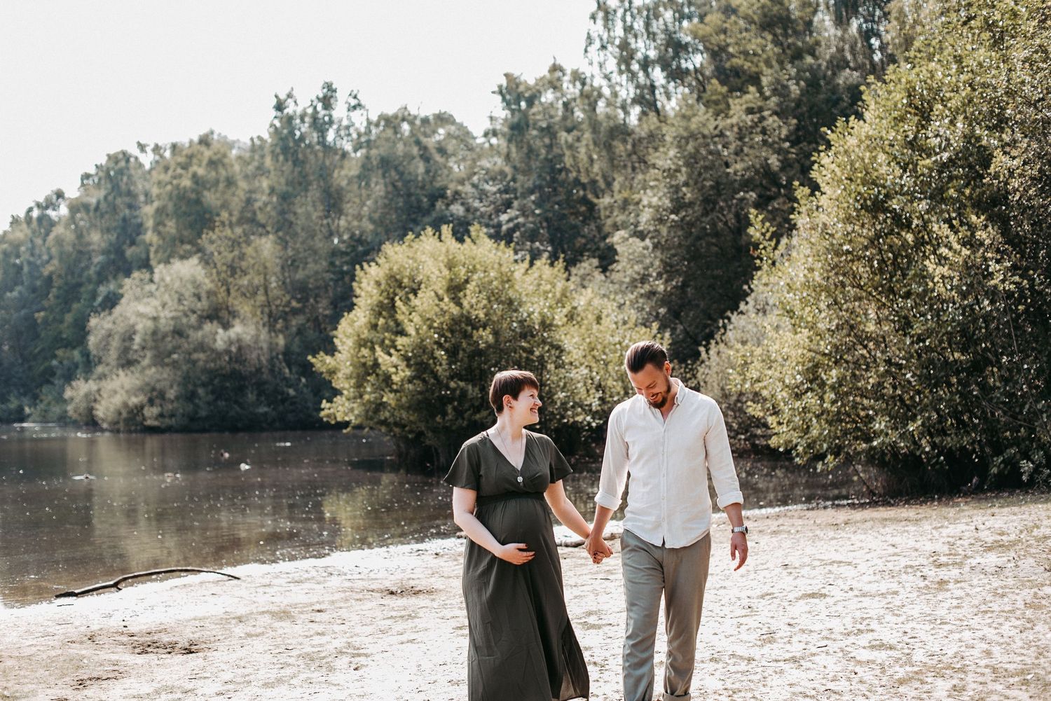 Ein Paar spaziert Hand in Hand am Ufer eines Sees entlang, umgeben von üppigem Grün und Bäumen an einem sonnigen Tag.