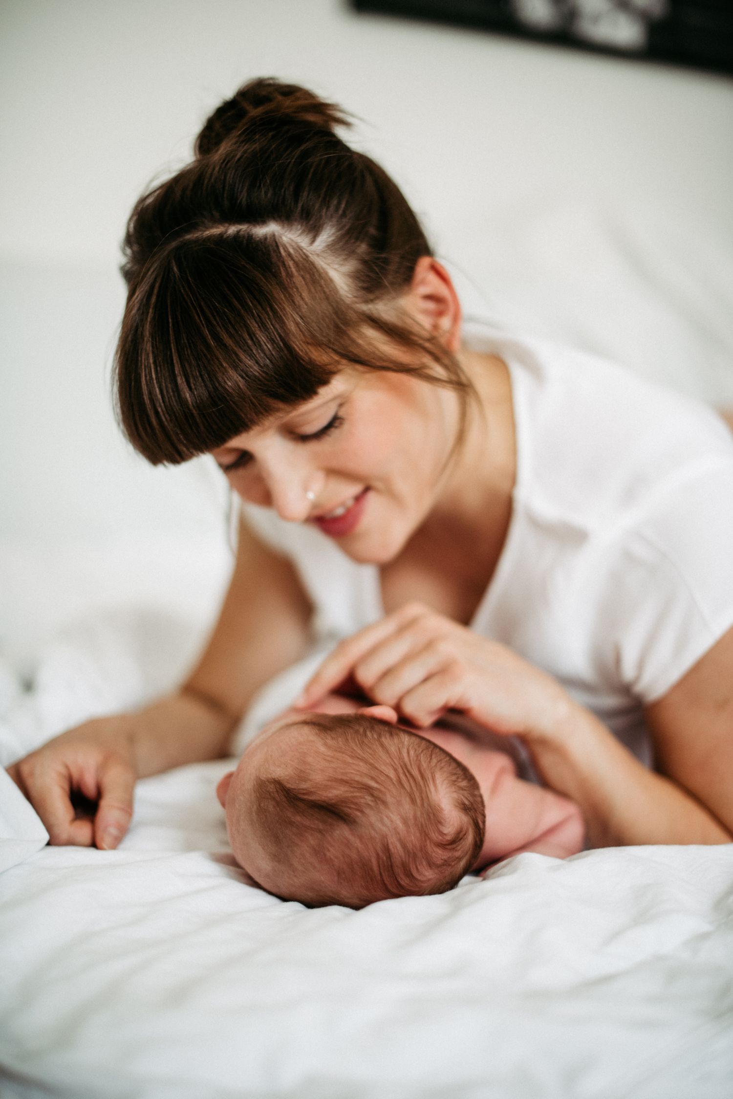 Frau mit braunen Haaren und Pony lächelt ein Baby an, das auf einem weißen Bett liegt. Beide wirken entspannt und glücklich.