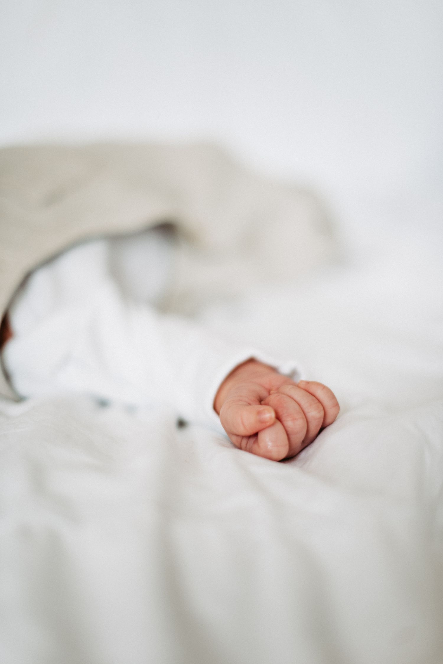 Nahaufnahme einer Babyhand, die auf einem weißen Bettlaken ruht. Die Hand ist leicht geballt und teilweise von einer Decke bedeckt.