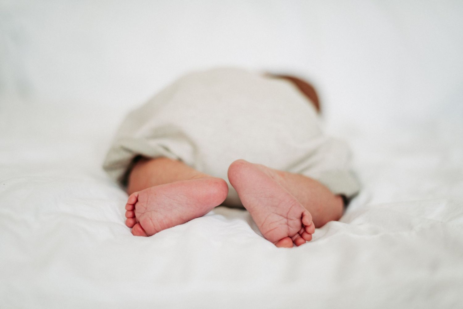 Ein Baby liegt auf dem Bauch auf einem weißen Bett, die Füße sind sichtbar. Es trägt eine helle Windel oder Hose.