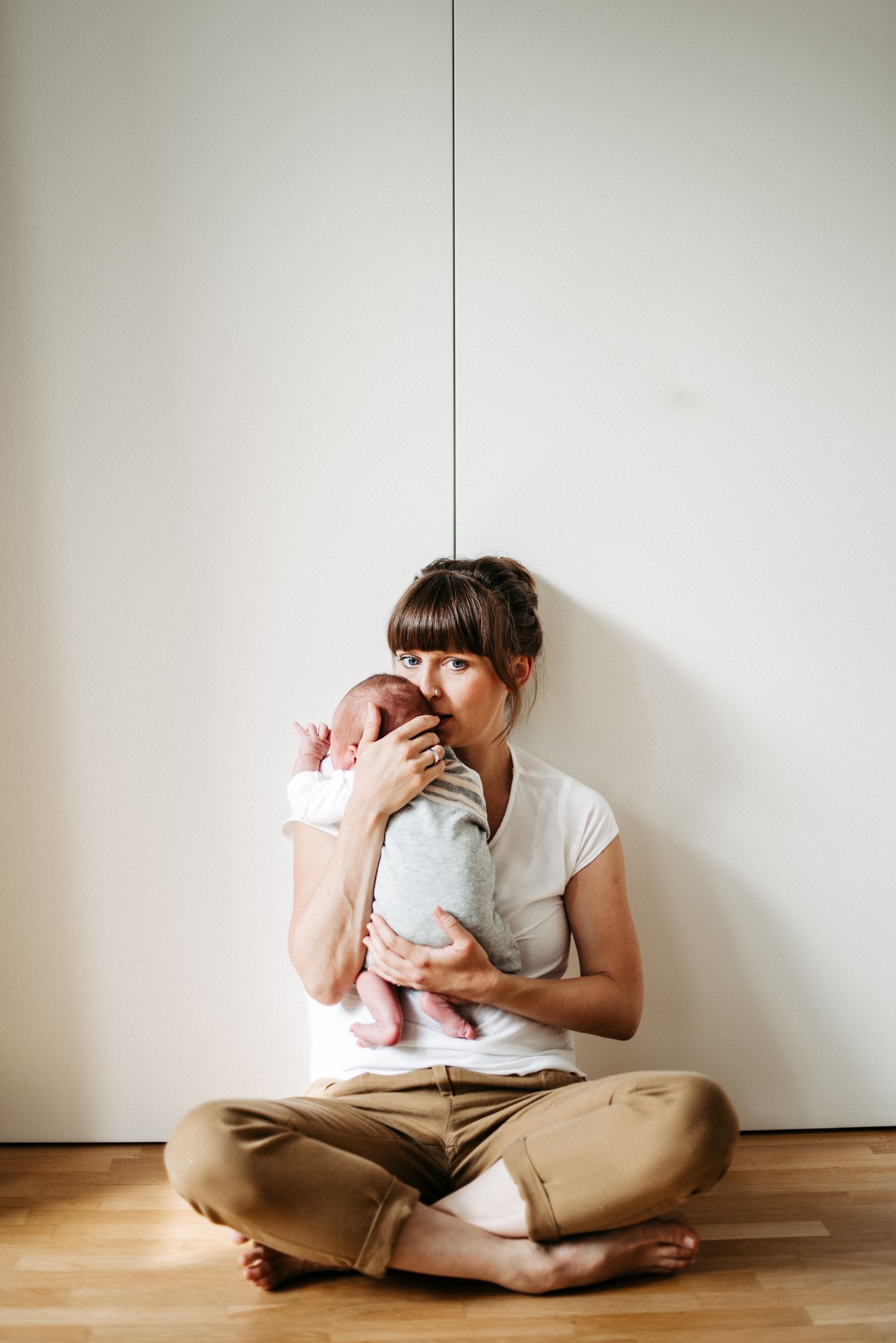 Frau sitzt auf Holzboden und hält ein Baby im Arm. Sie trägt ein weißes T-Shirt und beige Hose, das Baby ist in Grau gekleidet.