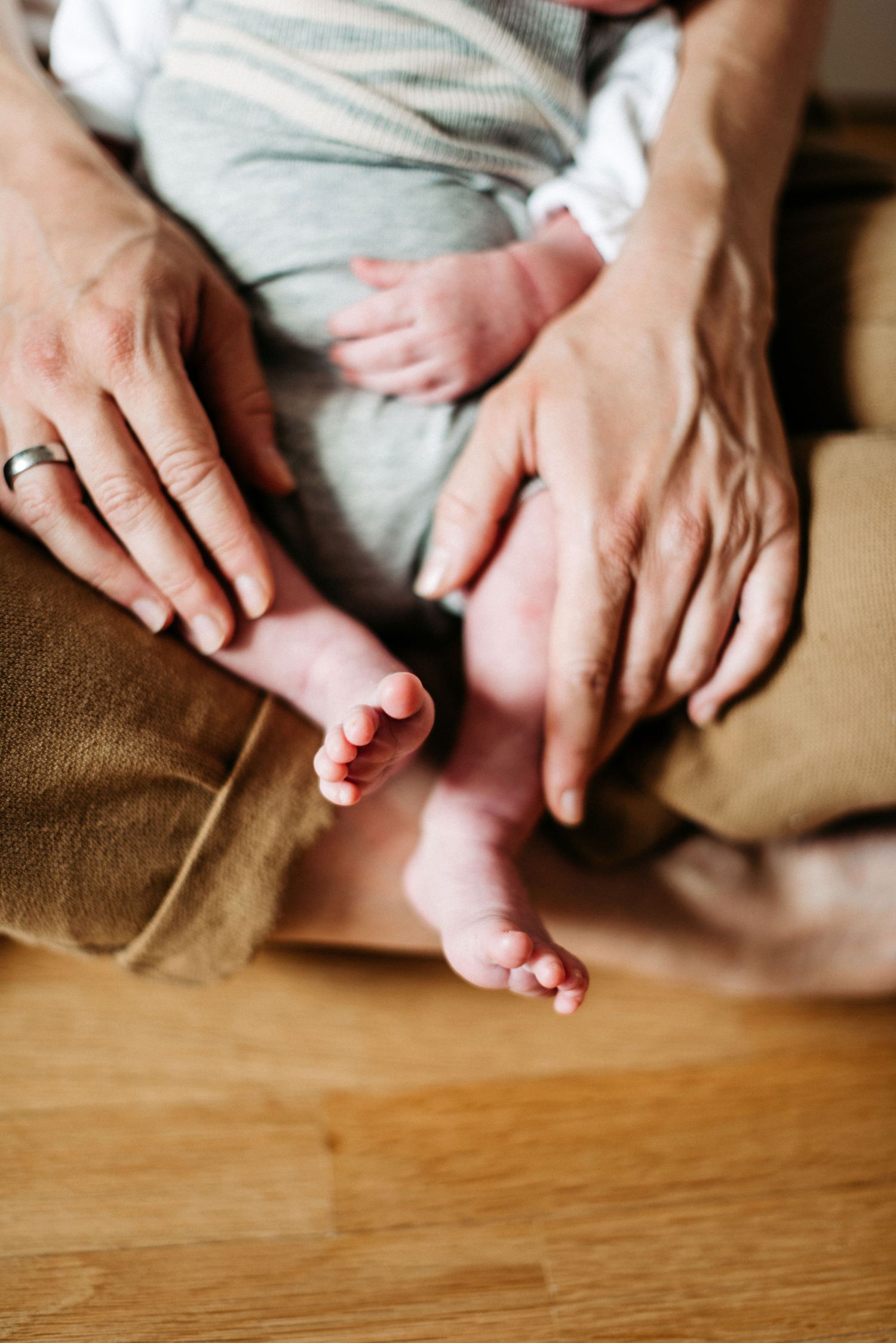 Erwachsene Hände halten sanft die Füße eines Babys, das auf einem Holzboden sitzt. Das Baby trägt einen gestreiften Strampler.