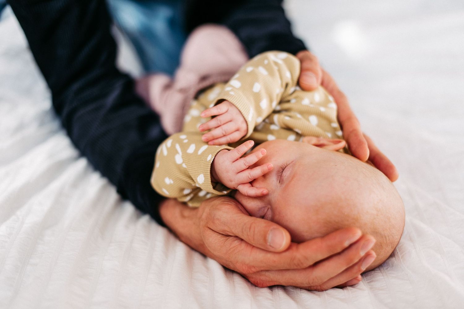 Ein Baby liegt in den Händen seines Vaters und hält eine Hand vor das GEsicht.