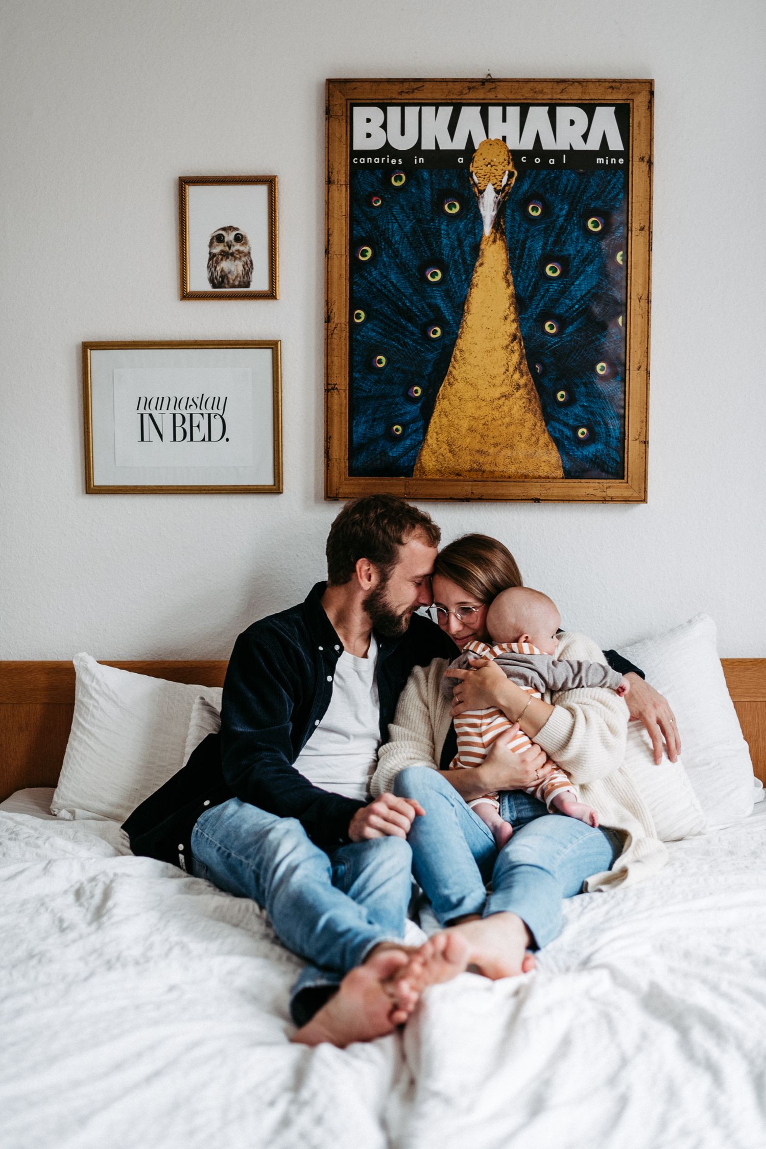 Ein Paar sitzt mit einem Baby auf einem Bett, umgeben von weißen Kissen. An der Wand hängt ein buntes Pfauenbild.