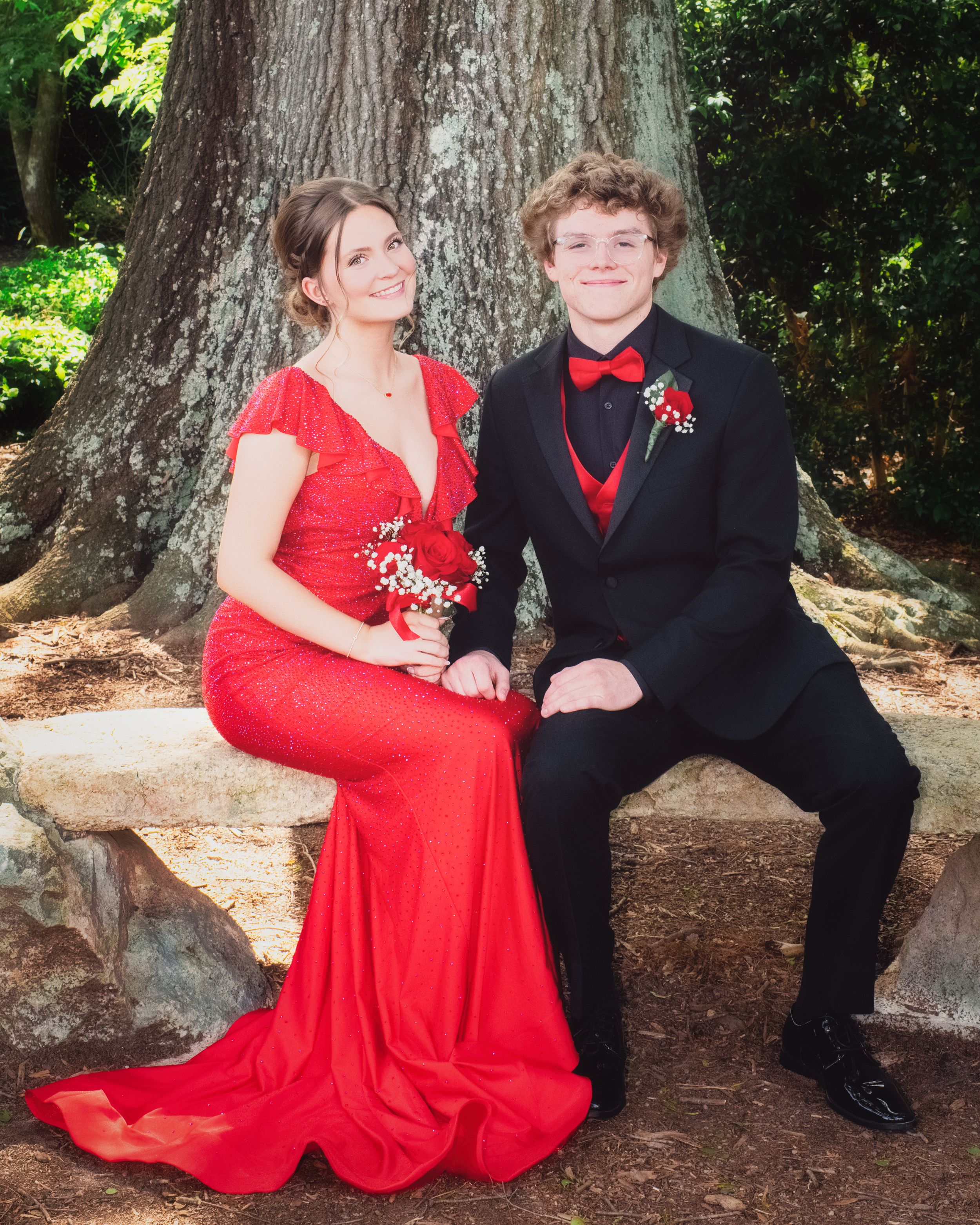 A couple in formal attire poses for prom photos, with her in a red gown and him in a black tuxedo by a large tree.
