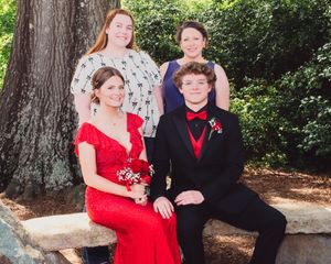 Formal group in elegant attire posed on stone bench outdoors with large tree trunk and greenery in background.