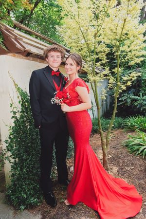 Couple posing in garden for prom photos with woman in red mermaid dress and man in black tuxedo.