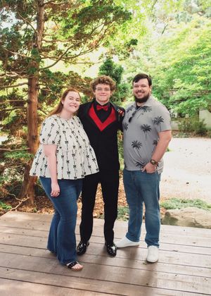 Three people stand together on a wooden deck surrounded by green trees on a sunny day.