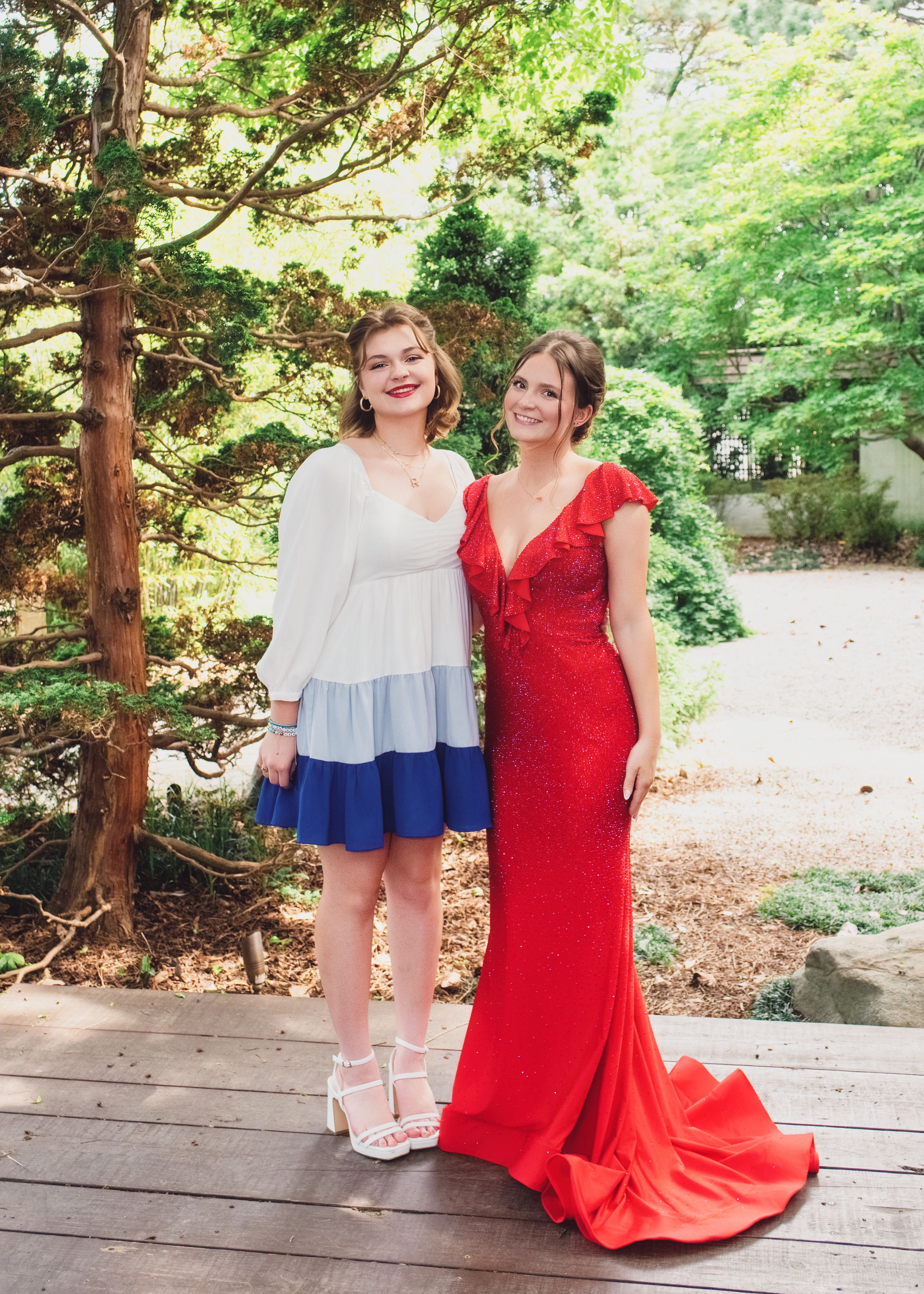 Two people in contrasting dresses stand on a wooden deck surrounded by lush greenery and trees.