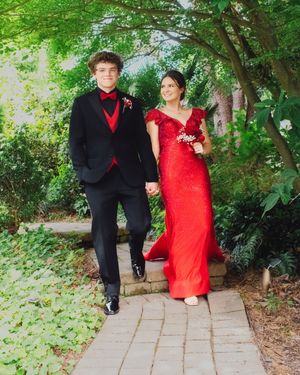 A couple in formal prom attire pose on a garden path with lush greenery, wearing a black suit and red gown.