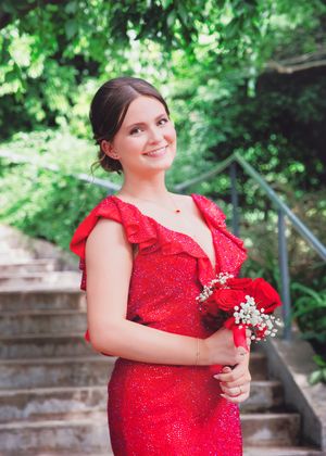 A person in a sparkly red formal dress holds a flower bouquet while posing on outdoor stone steps.