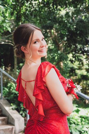 Sparkling red formal dress with ruffled back detail and delicate beading in an outdoor garden setting.