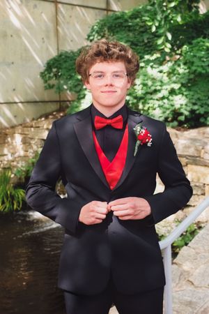 Formal portrait of someone in a black tuxedo with red bow tie and vest against a stone wall and garden backdrop.