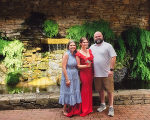 Three people pose together in front of a decorative stone waterfall wall with cascading greenery.