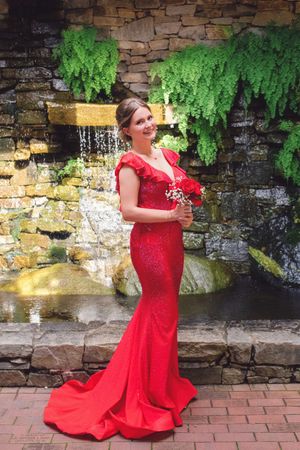 A person in a stunning red mermaid gown poses elegantly by a stone waterfall feature.