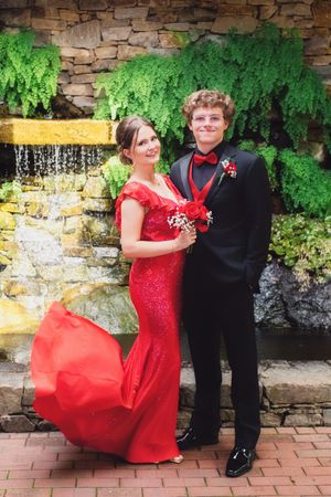 A formal couple poses in front of a stone wall with greenery, wearing red gown and black tuxedo for prom or formal event.