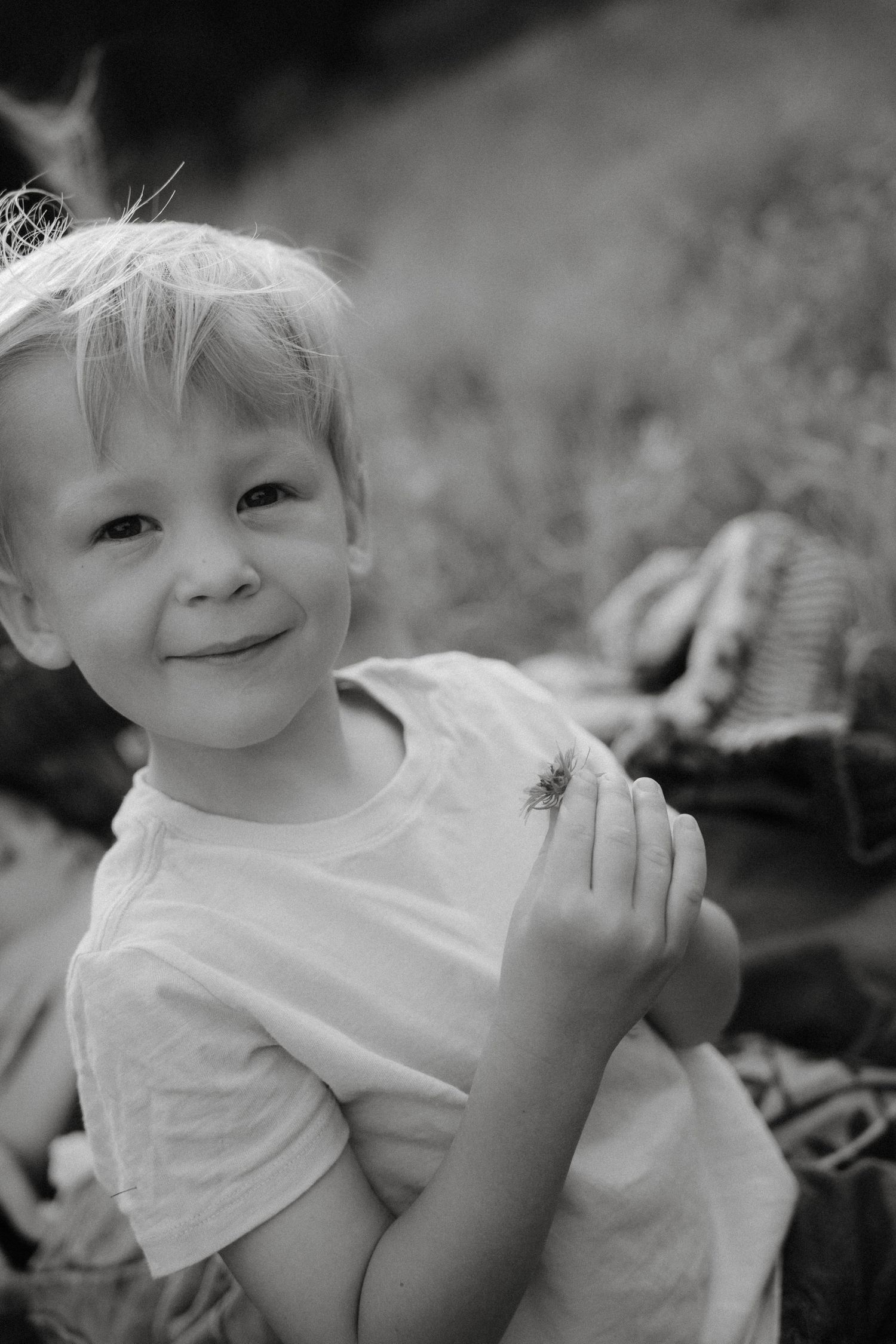 Black and white portrait of a child in a white t-shirt smiling at the camera outdoors.