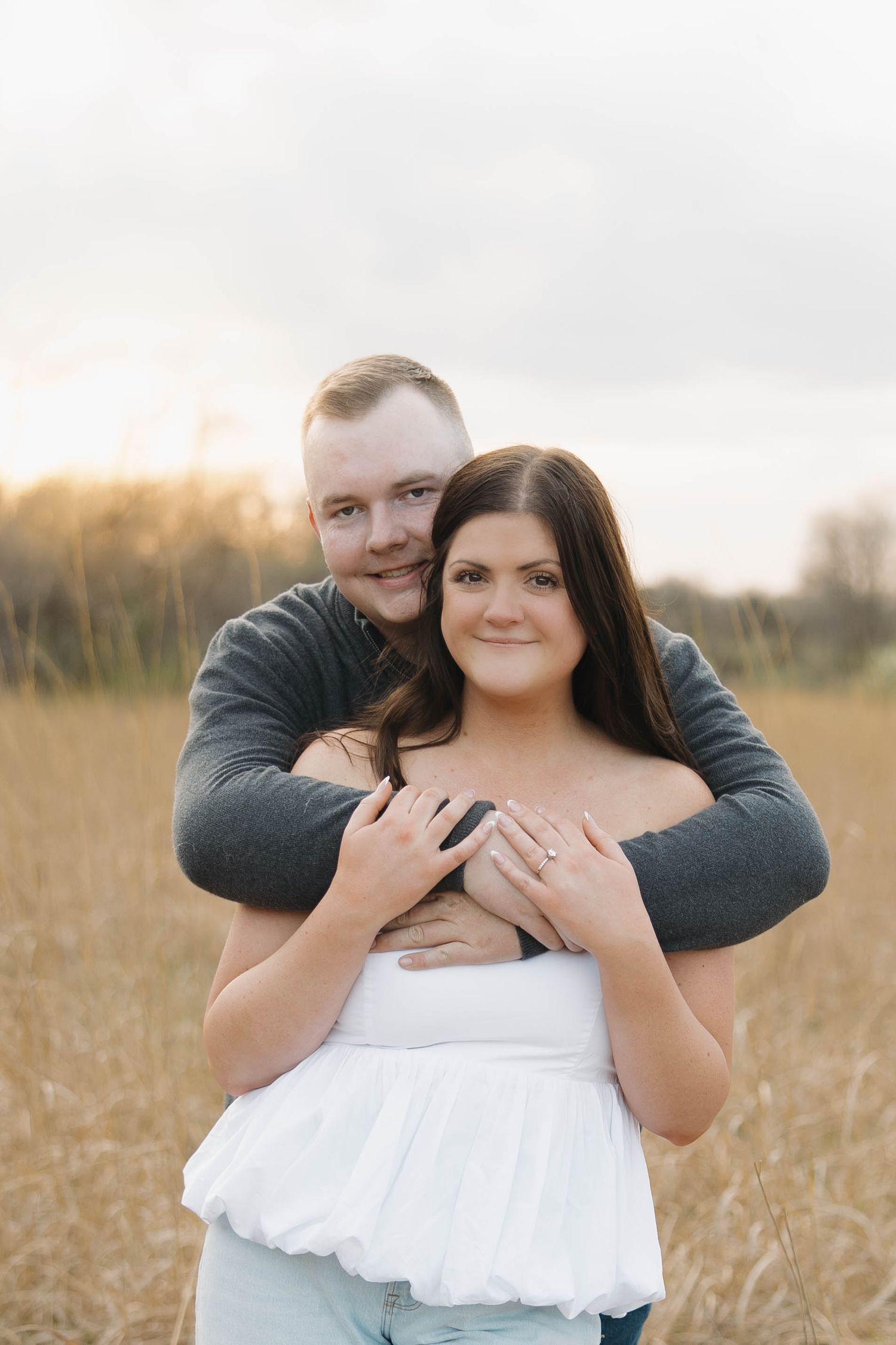 A romantic couple embraces in a golden wheat field at sunset while wearing casual attire.