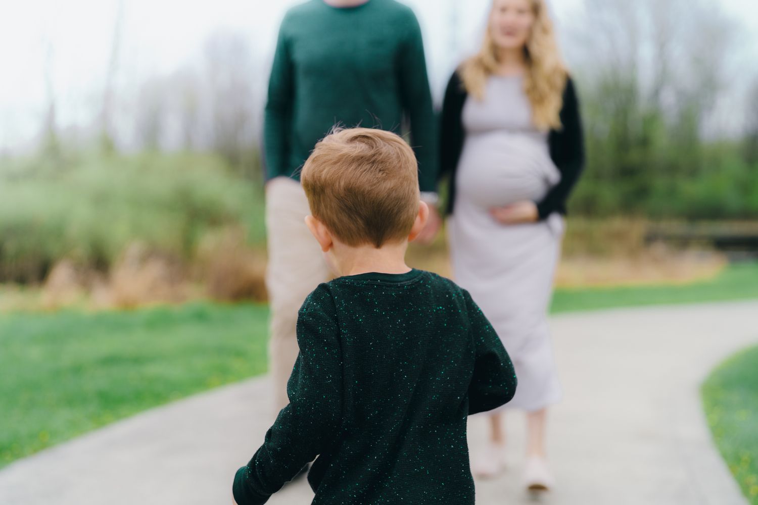 Young child in dark clothing runs towards blurred figures standing on a path in a garden setting.