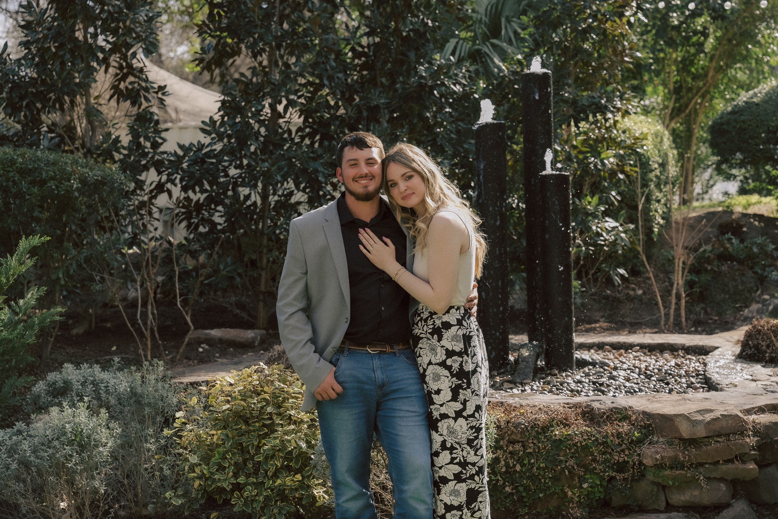 A couple poses together in a garden with green foliage and stone landscaping as their backdrop.