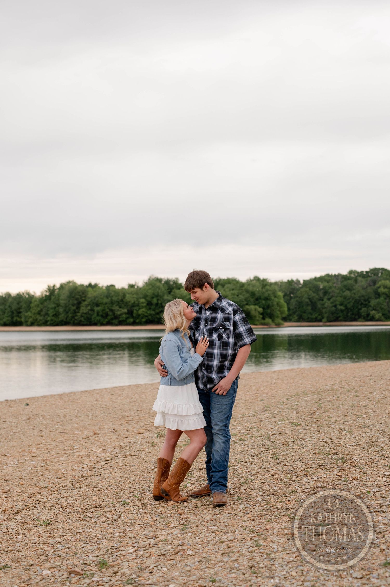 A couple shares a romantic moment on a beach by a lake surrounded by trees during an overcast day.