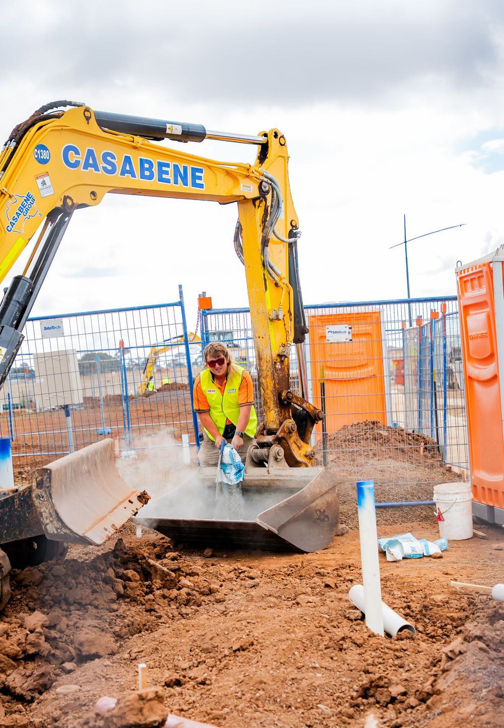 Construction worker in safety gear operates yellow Casabene excavator while installing underground utilities at jobsite.